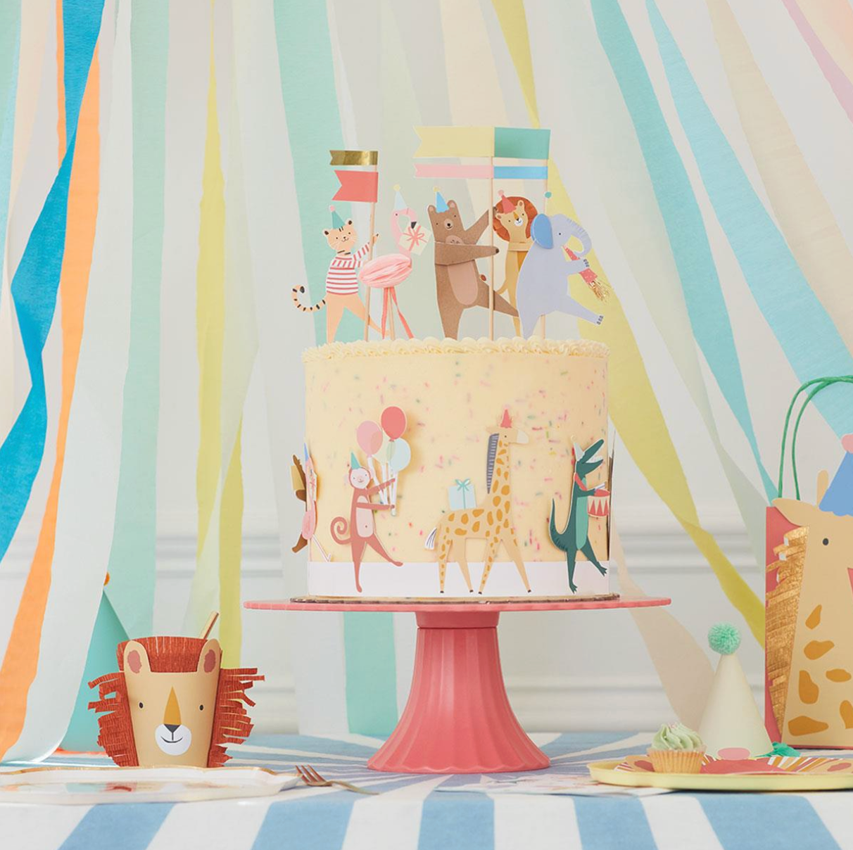 Children's birthday cake with animal-themed decorations on a pink stand against a colorful striped curtain.