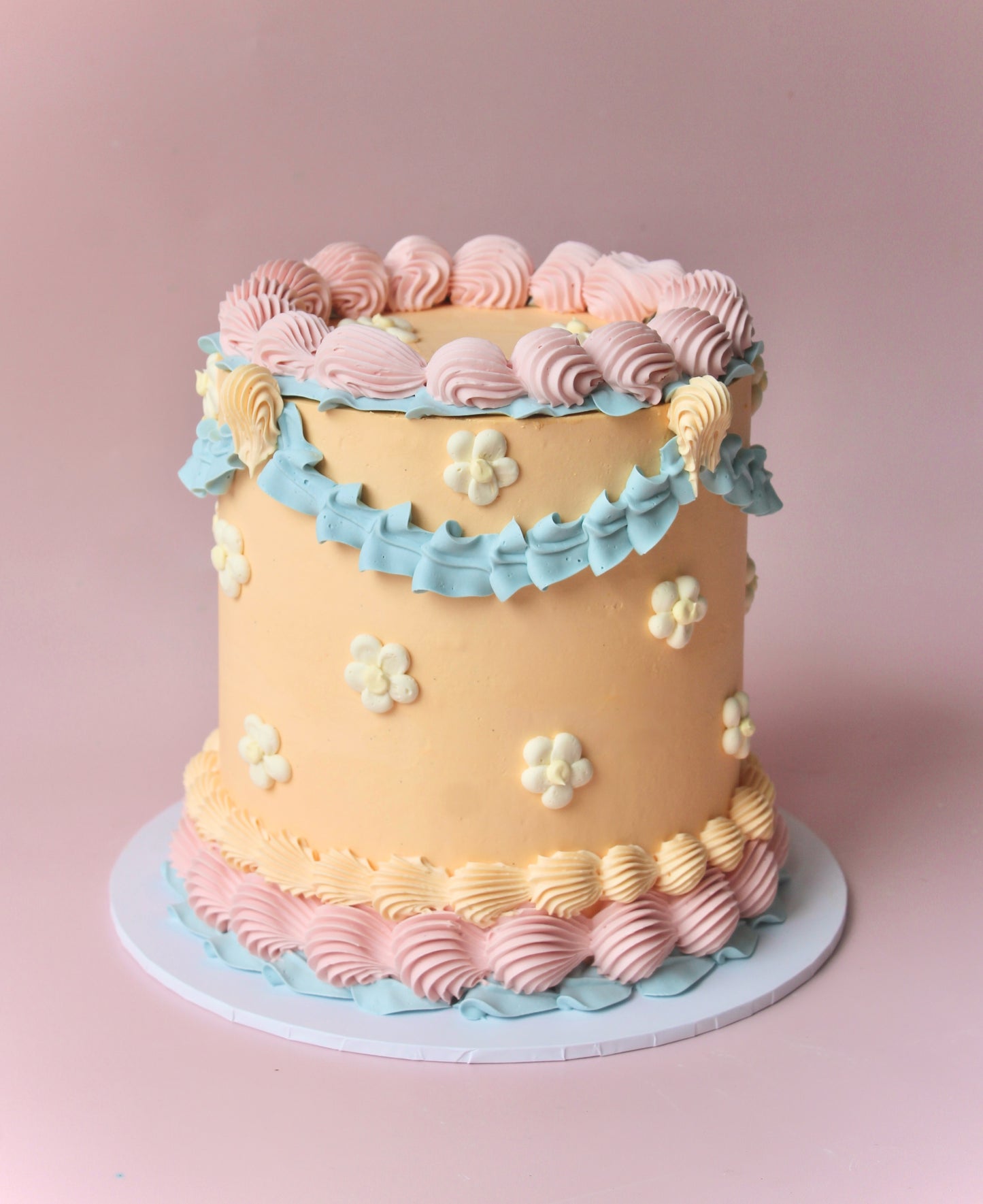 A multi-layered cake with beige, blue, and pink icing, decorated with white flowers andlemon yellow centers