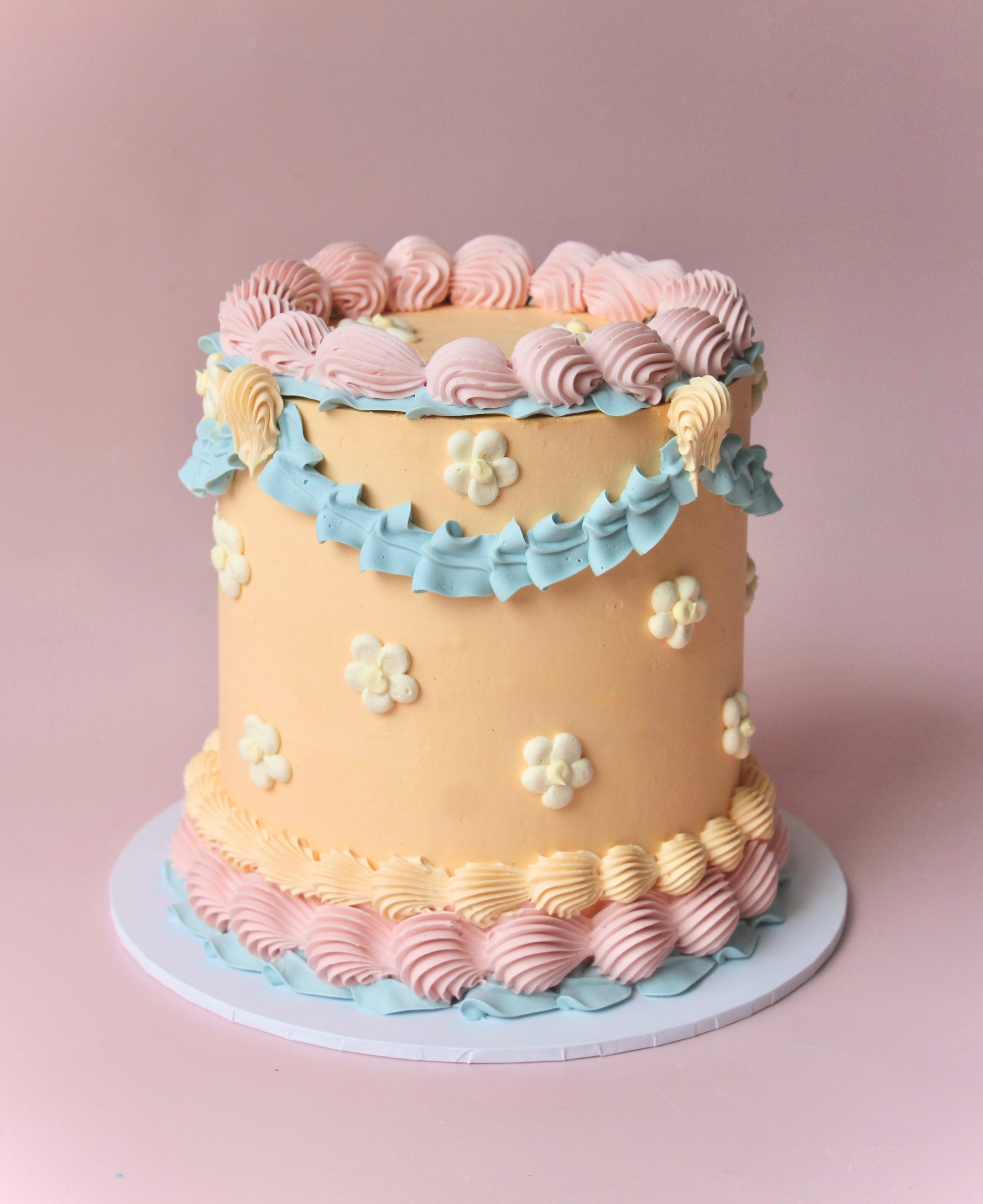 A multi-layered cake with beige, blue, and pink icing, decorated with white flowers andlemon yellow centers